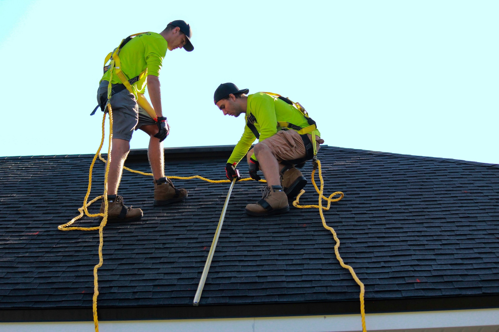 Two roofers in safety harnesses measuring on a dark asphalt shingle roof under a clear blue sky